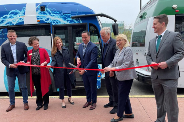 A group of seven people are cutting a red ribbon in front of two buses, one blue and one green, during an outdoor ceremony.