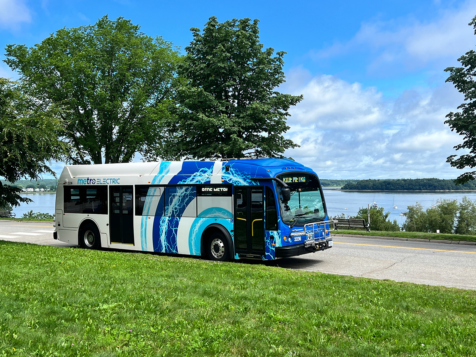 Electric bus parked by a road, with trees and water in the background.