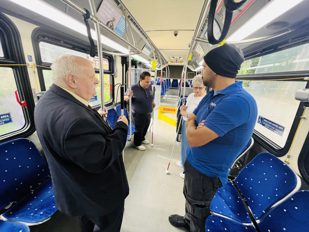 Group of people talking inside a city bus, some holding white canes. The seats are blue with white polka dots, and the floor is gray.