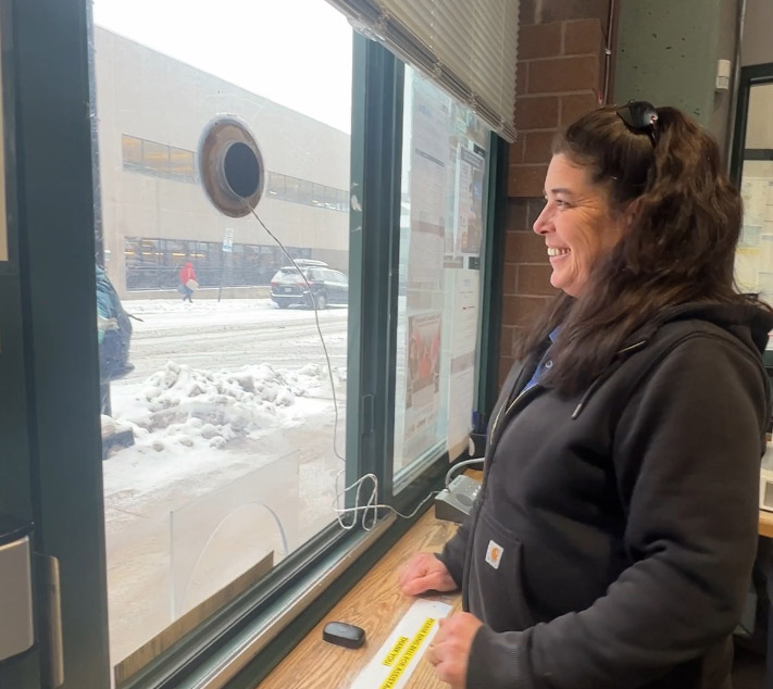 A woman in a black hoodie stands inside, smiling and looking out a window. Snow is visible outside the large window.