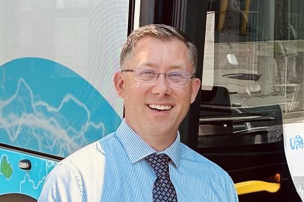 Man in glasses and a blue shirt stands smiling in front of a bus.