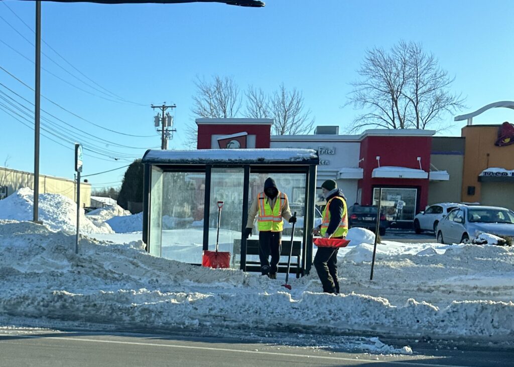 Two people in reflective vests shoveling show out of a bus stop with a bus shelter