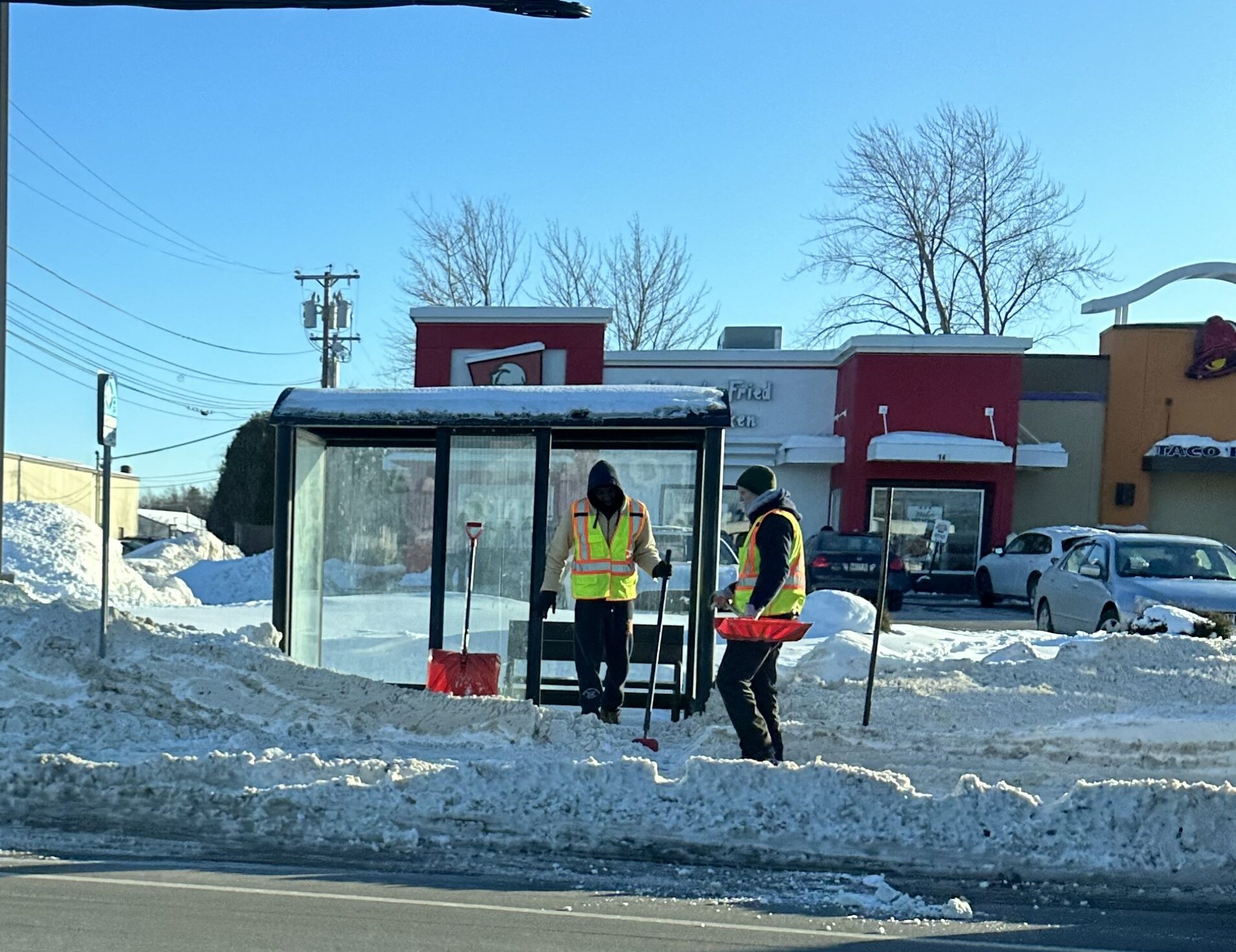 Two people in reflective vests shoveling show out of a bus stop with a bus shelter